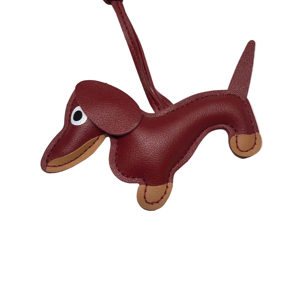 A dachshund-shaped leather bag charm or luggage tag in dark red with tan accents, featuring a white and black eye detail and a matching red strap, photographed on a light gray surface.