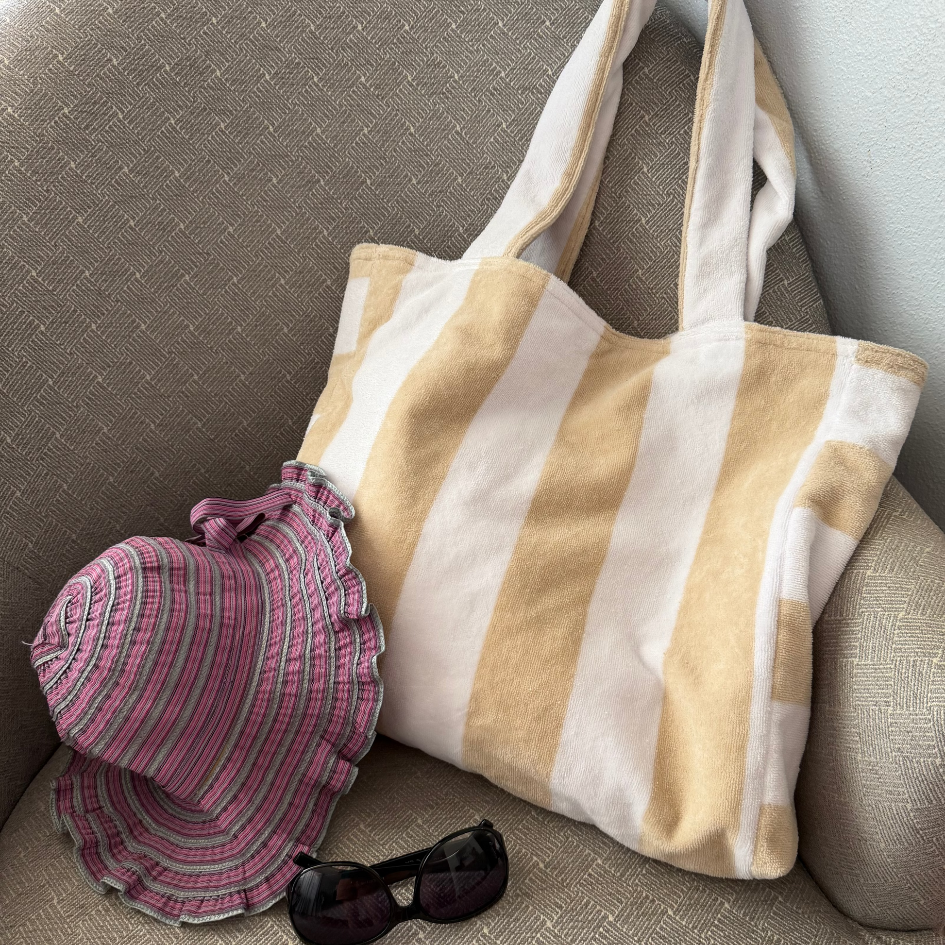 A striped canvas tote bag in beige and white sits on a gray upholstered armchair, styled with a pink striped sun hat and black sunglasses.